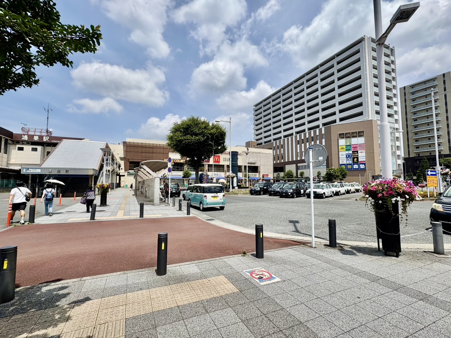 鴻巣駅東口の駅前広場 - まつげエクステサロン「アイデザイン ツツ.」への道順案内、駅舎とタクシー乗り場、商業施設が見える明るい駅前エリア