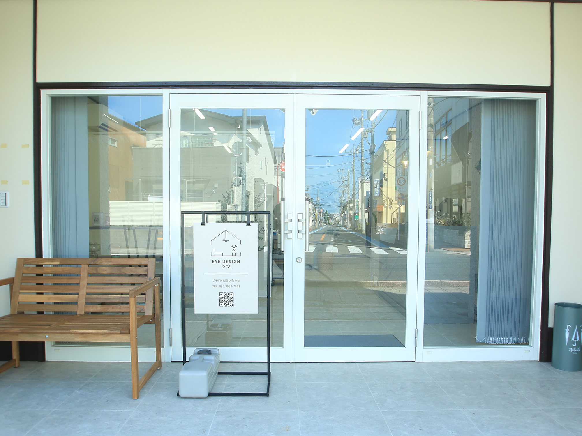 アイデザイン ツツ.店舗外観 - 鴻巣市のまつげエクステ専門サロン最終目的地、クリーム色の建物に大きなガラス窓、白い看板にEYE DESIGN ツツ.のロゴ、木製ベンチ設置、鴻巣駅東口徒歩5分
